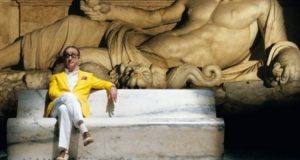 Toni Servillo con la giacca gialla seduto ai piedi di una statua colossale