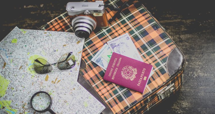 Valigia con, sopra, un paio di occhiali da sole, una macchina fotografica, una cartina, un passaporto e delle banconote